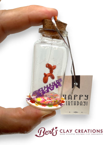 Small glass bottle with a cork lid containing a colorful clay ornament, held by a hand. 'Happy Birthday' card in the background with 'Bert's Clay Creations' branding.